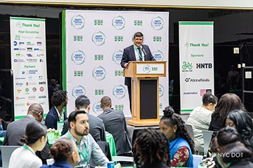 A man in formal attire at a podium, addressing an audience with his remarks. Behind the man, is a backdrop with logos of the event, sponsors, and partners.